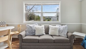 Un salon avec un canapé gris, deux coussins blancs et moelleux, une table à manger en bois avec des chaises, et une fenêtre donnant sur une maison et des arbres.