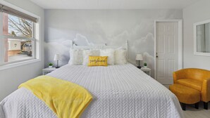 Een slaapkamer met een groot bed, een gele plaid, een wit hoofdeinde en een geel kussen met de tekst 'sunshine'.