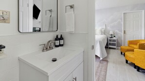 A modern bathroom with a white sink, mirror, and towel rack. There is a bed with a white blanket and a nightstand with a lamp. - The Luxe King Room 5 / Pleasant Valley Arcadia Resort (Arcadia)