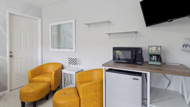 A compact room with a white door, two yellow armchairs, a small white table, a microwave, a coffee machine, and a flat-screen TV mounted on the wall. - The Luxe King Room 5 / Pleasant Valley Arcadia Resort (Arcadia)