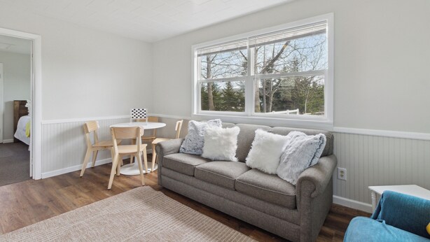 A living room with a grey sofa, two wooden chairs, a small round table, and a large window with blinds.