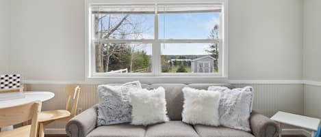 A living room with a gray sofa, two white fluffy pillows, a wooden dining table with chairs, and a window with a view of a house and trees.