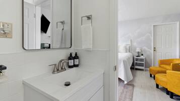 A modern bathroom with a white sink, mirror, and towel rack. There is a bed with a white blanket and a nightstand with a lamp.