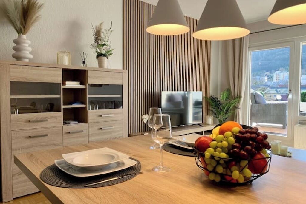 A modern dining area with a wooden table, a fruit bowl, and a television in the background.