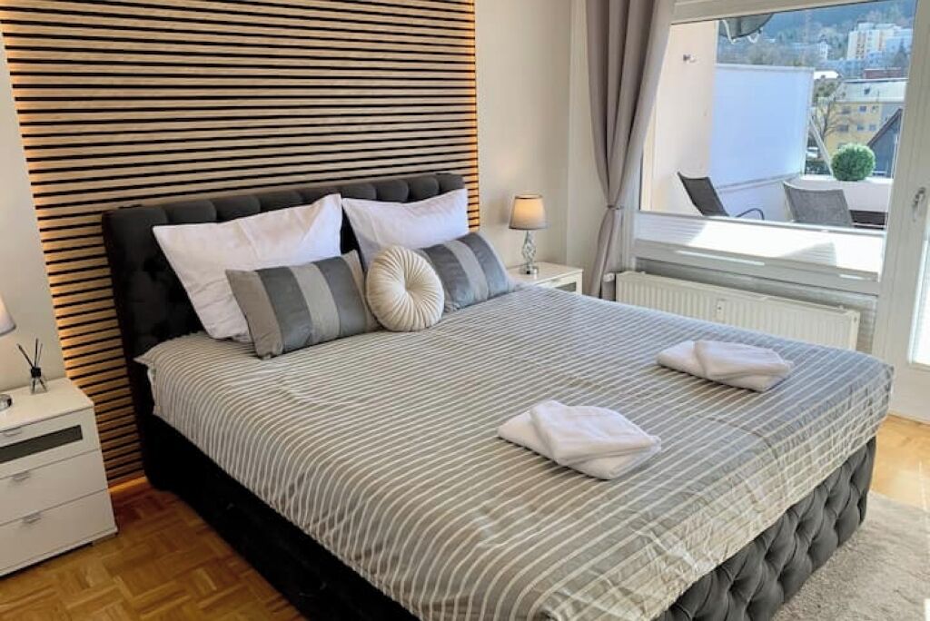 Ein ordentlich bezogenes Bett mit Streifenmuster, ein schwarzer, möhrner Kopfteil und ein Fenster mit Blick auf Gebäude und Grünflächen.