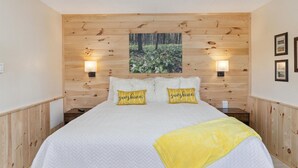 A bedroom with a wooden headboard, two bedside lamps, a bed with white bedding and a yellow throw, and a forest-themed wall art.