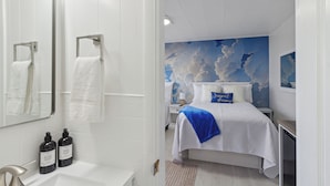 A hotel room with a white bathroom, a bed with a blue pillow, and a cloud mural on the wall. - Deluxe Double Studio 6 (Arcadia)