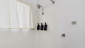 A bathroom with white tiled walls, a showerhead, and three black bottles on the wall. - Deluxe Double Studio 6 (Arcadia)