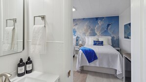 Quarto de hotel com banheiro branco, cama com travesseiro azul e um mural com tema de nuvens na parede.