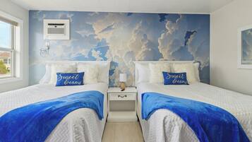 A bedroom with two beds, each with a pillow labeled "Sweet dreams," a nightstand with a lamp, and a wall mural of clouds.