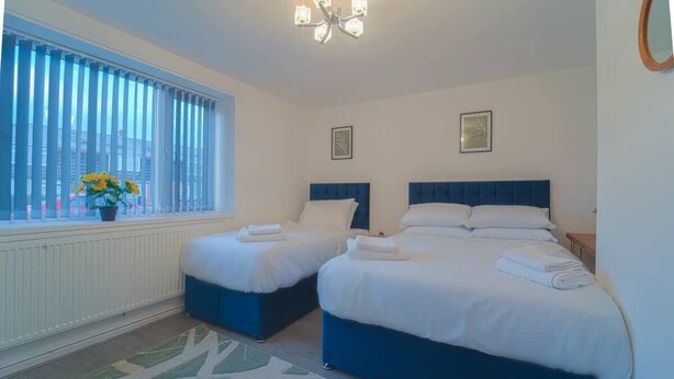 A hotel room with two beds, a window with blinds, a radiator, a chandelier, and a view of buildings outside.