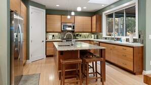 Une cuisine moderne avec des meubles en bois, un réfrigérateur en acier inoxydable, un four à micro-ondes et une grande fenêtre donnant sur une forêt.