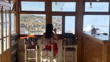 Habitación con vistas a un pueblo a través de amplios ventanales, muebles de madera y una cesta tejida.
