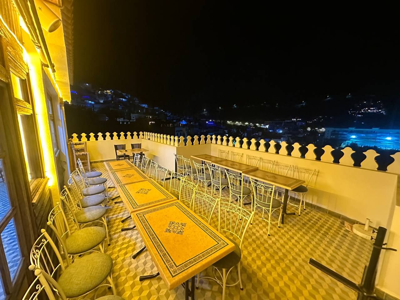 Una terraza en la azotea con mesas y sillas, vista nocturna de la ciudad y barandilla con detalles decorativos.