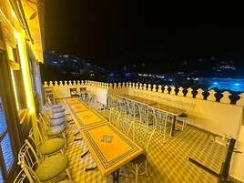 Una terraza en la azotea con mesas y sillas, vista nocturna de la ciudad y barandilla con detalles decorativos.