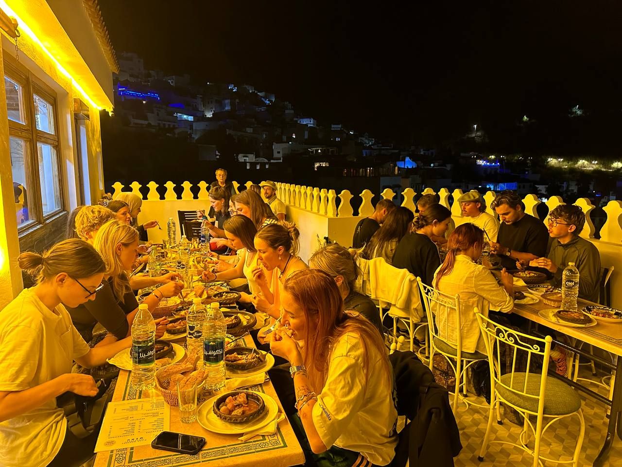 A group of people dining at an outdoor restaurant with tables arranged in rows, overlooking a hillside.