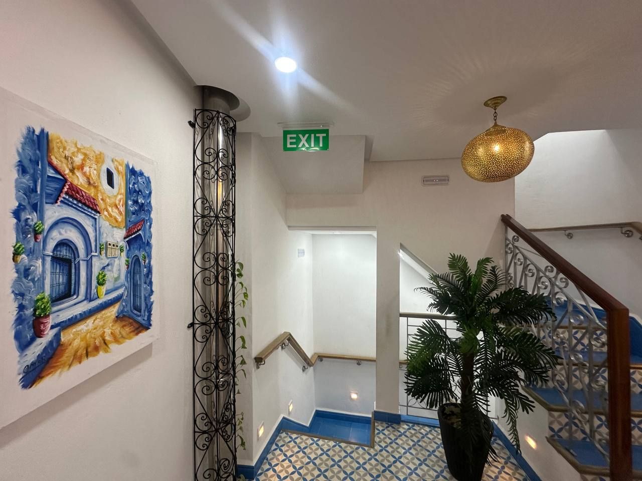 Pasillo de hotel con letrero de salida, una escalera con escalones azules, una planta en maceta y un cuadro decorativo en la pared.