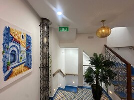Pasillo de hotel con letrero de salida, una escalera con escalones azules, una planta en maceta y un cuadro decorativo en la pared.