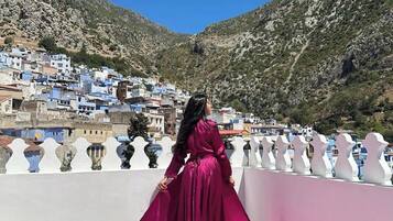 Una persona con un vestido rosa largo parada en un balcón que domina un pueblo montañoso de edificios blancos.
