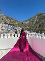 Una persona con un vestido rosa largo parada en un balcón que domina un pueblo montañoso de edificios blancos.