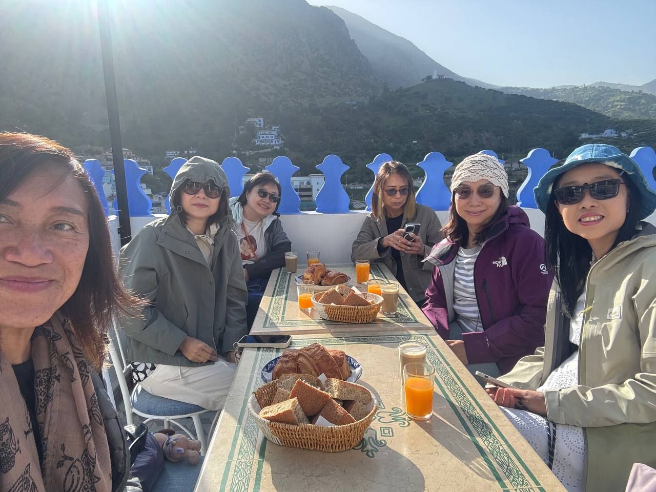 A group of people seated at a table with food and drinks, overlooking a mountainous landscape.