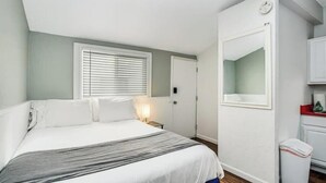 A bedroom with a bed, a bedside table, a window with blinds, a door, and a small trash bin. - San Diego Surf Shack #3 -1/2 Block to the Pacific! (San Diego)