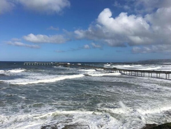 Escena costera con un muelle que se adentra en el mar, olas rompiendo contra la orilla y un cielo parcialmente nublado.