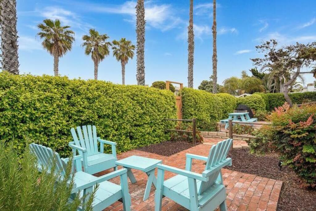 A patio area with light blue Adirondack chairs and a picnic table, surrounded by a well-maintained hedge and palm trees.