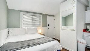 Une chambre à coucher comprenant un lit, une table de chevet, une fenêtre avec des stores, une porte et un petit meuble.