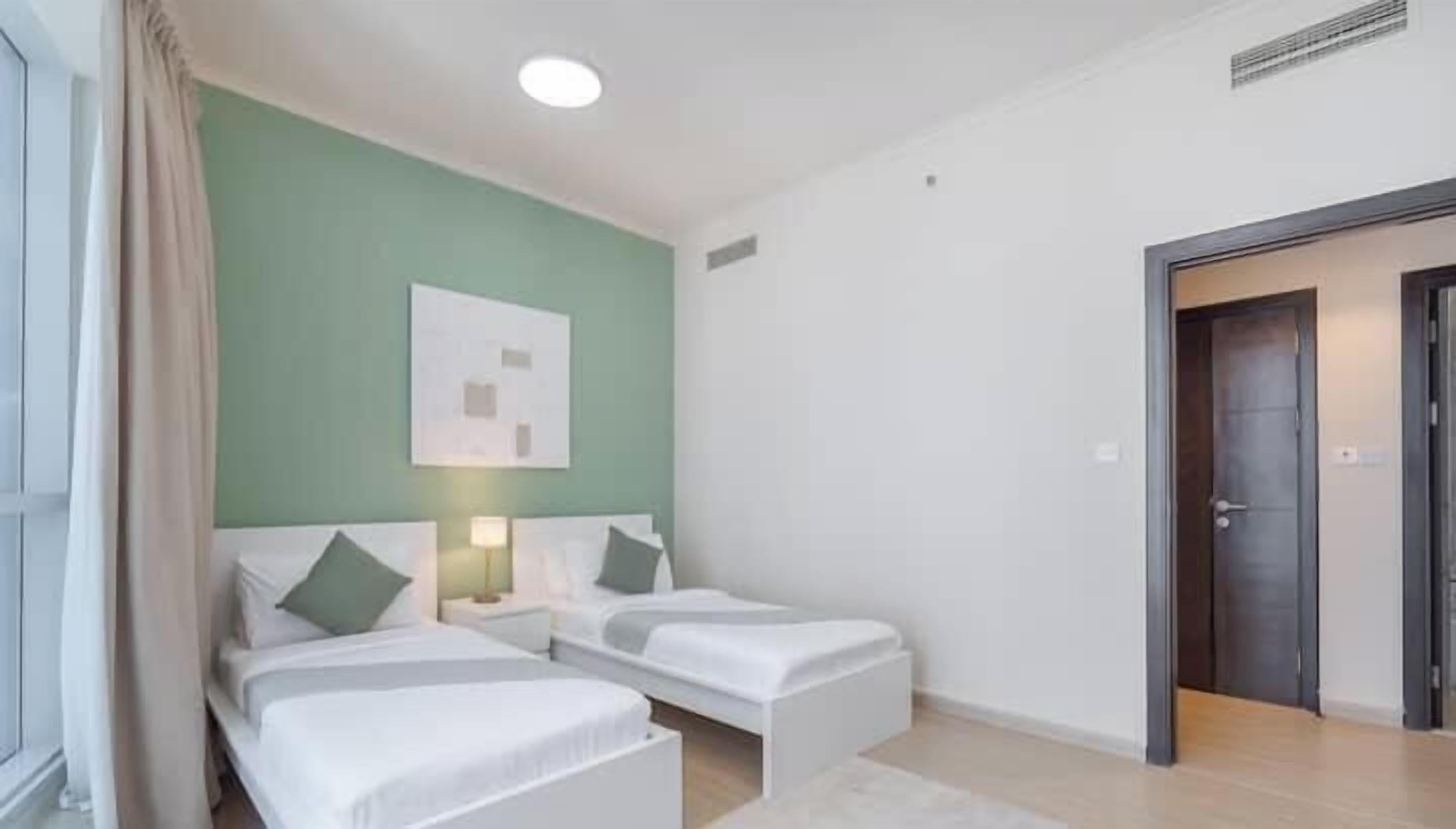 A hotel room with two beds, a green wall, a white ceiling, and a door to the right.
