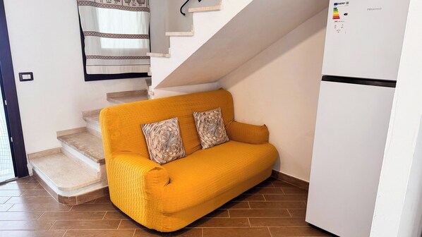 A yellow sofa with woven cushions in a corner of a room with a white refrigerator and a staircase.