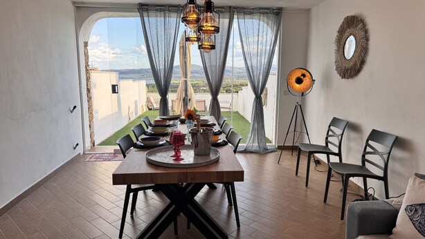 A dining area with a table set for four, a floor lamp, and a view of the landscape through large windows.