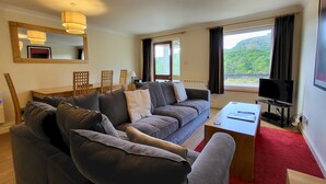 A modern living room with a grey sofa, a wooden coffee table, and a TV. There is a large window with a view of greenery and mountains. - Lingmell Fell View, Chapel Stile (Chapel Stile)