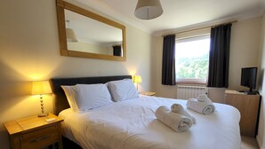 A neatly made bed with white linens and towels, a bedside lamp, a nightstand, a television, and a window with curtains. - Lingmell Fell View, Chapel Stile (Chapel Stile)