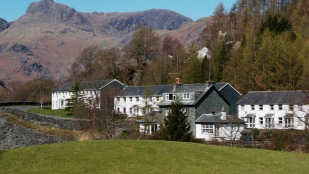 A large, white building with multiple chimneys, surrounded by a grassy area and trees, with mountains in the background. - Lingmell Fell View, Chapel Stile (Chapel Stile)