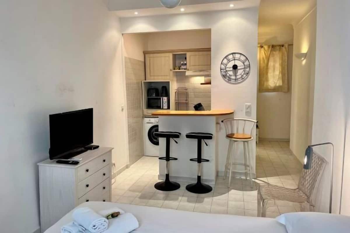 A compact hotel room with a bed, a TV, a desk with two stools, a kitchen area, and a clock on the wall.