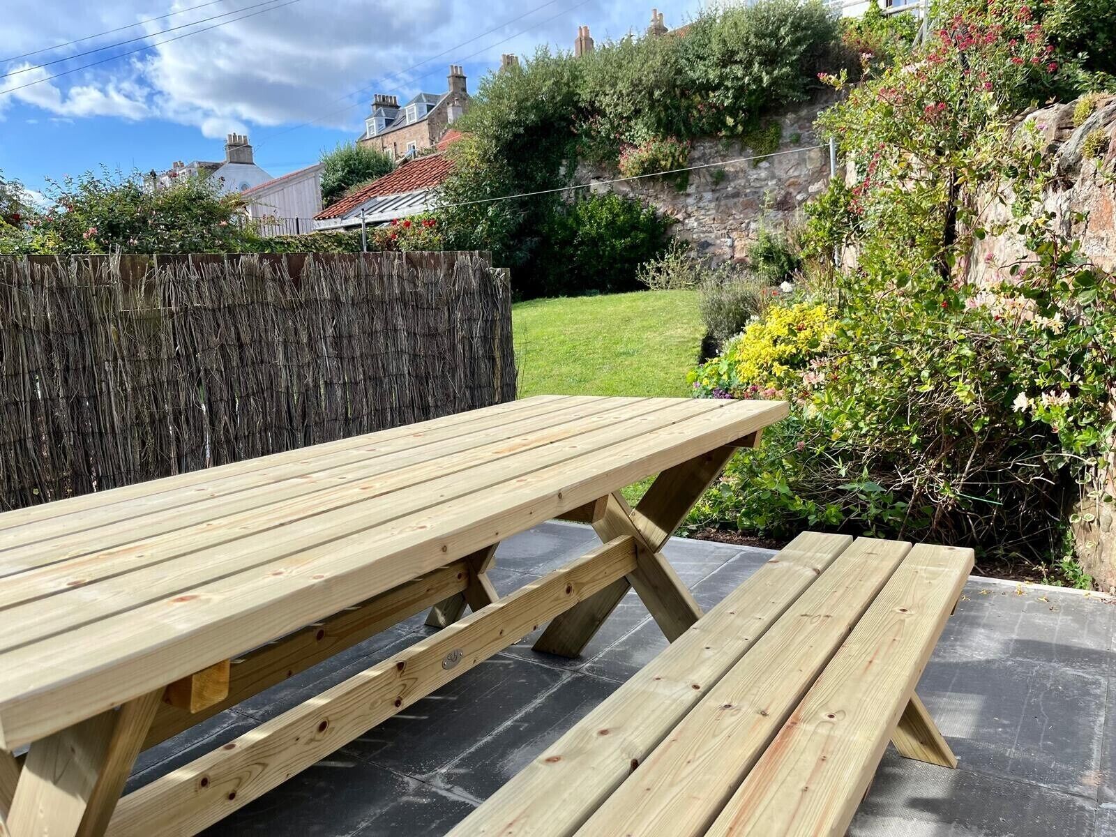 Une table de pique-nique en bois, avec des bancs, située dans un espace jardin doté d’un sol en pierre, entourée de verdure et d’un mur de pierre.