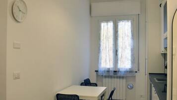 Una habitación pequeña y limpia, con una mesa y sillas blancas, una ventana con cortinas translúcidas y un reloj en la pared.
