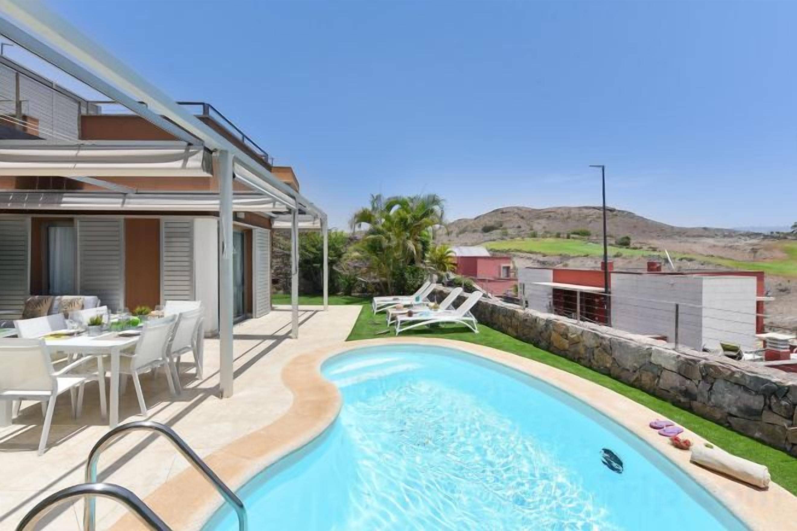 A swimming pool with a clear blue water, surrounded by a patio area with white chairs and a table.