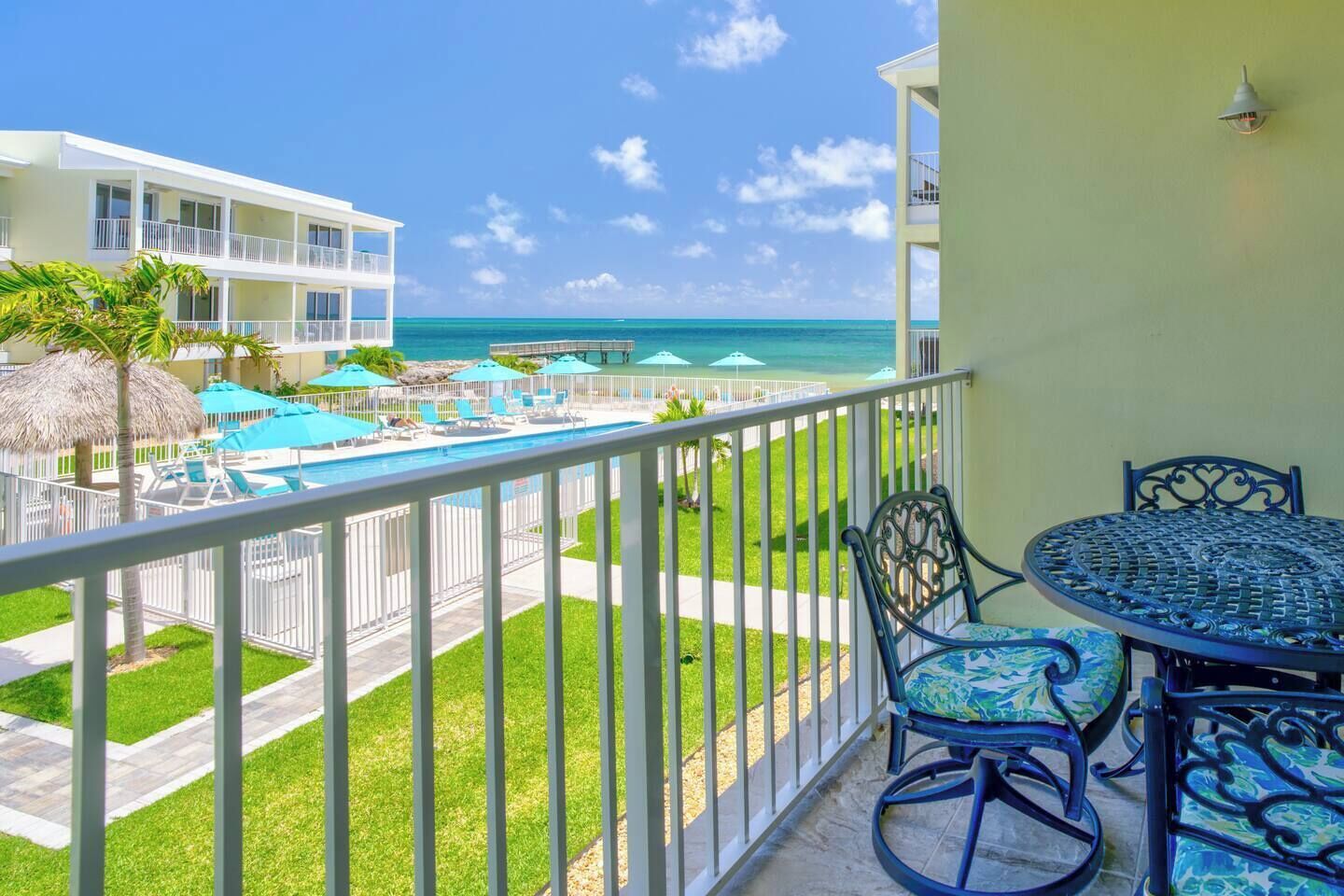 A balcony with a view of a pool and beach, a dining table with chairs, and a thatched umbrella.