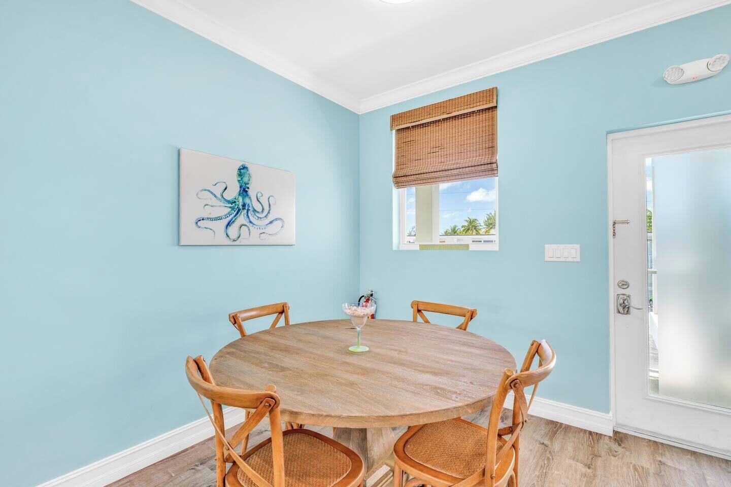 Een kamer met een ronde houten tafel en vier rieten stoelen, een schilderij van een octopus, een raam met een bamboe rolgordijn en een deur met een matglazen paneel.
