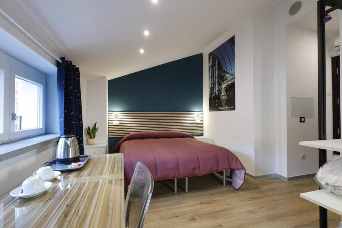 A modern hotel room with a bed, a dining table with a kettle, cups, and a chair.
