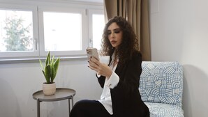A person sitting on a patterned sofa in a room with a potted plant on a small table, a window with curtains, and a plain wall. - New Pescara City b&b  (Pescara)