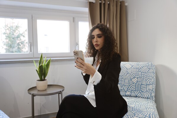 A person sitting on a patterned sofa in a room with a potted plant on a small table, a window with curtains, and a plain wall. - New Pescara City b&b  (Pescara)