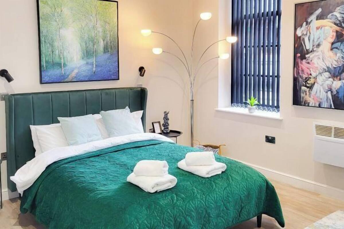 Une chambre d’hôtel avec une couvre-lit verte, des oreillers blancs, un tableau représentant une forêt, un grand lampadaire et une fenêtre avec des stores.