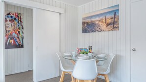 Un coin repas avec une table en verre, des chaises blanches et un tableau représentant une scène de plage.
