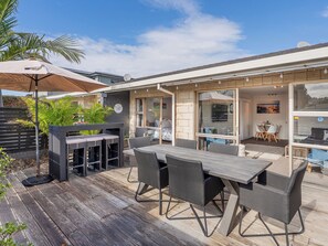 Een terras buiten met een houten dek, een eettafel met stoelen, een barbecue en een overdekt zitgedeelte met parasols.