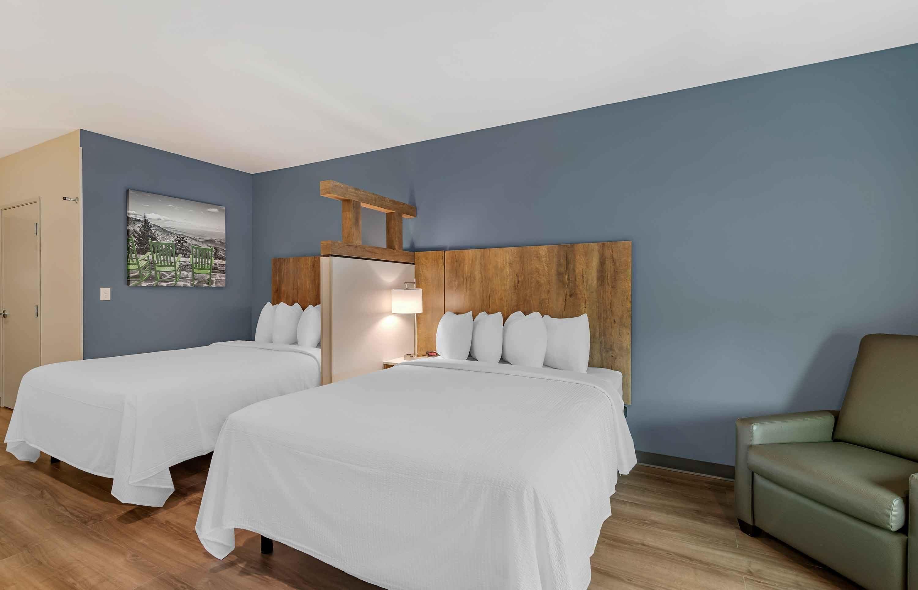 A hotel room with two beds, a wooden headboard, a green chair, and a painting on the wall.