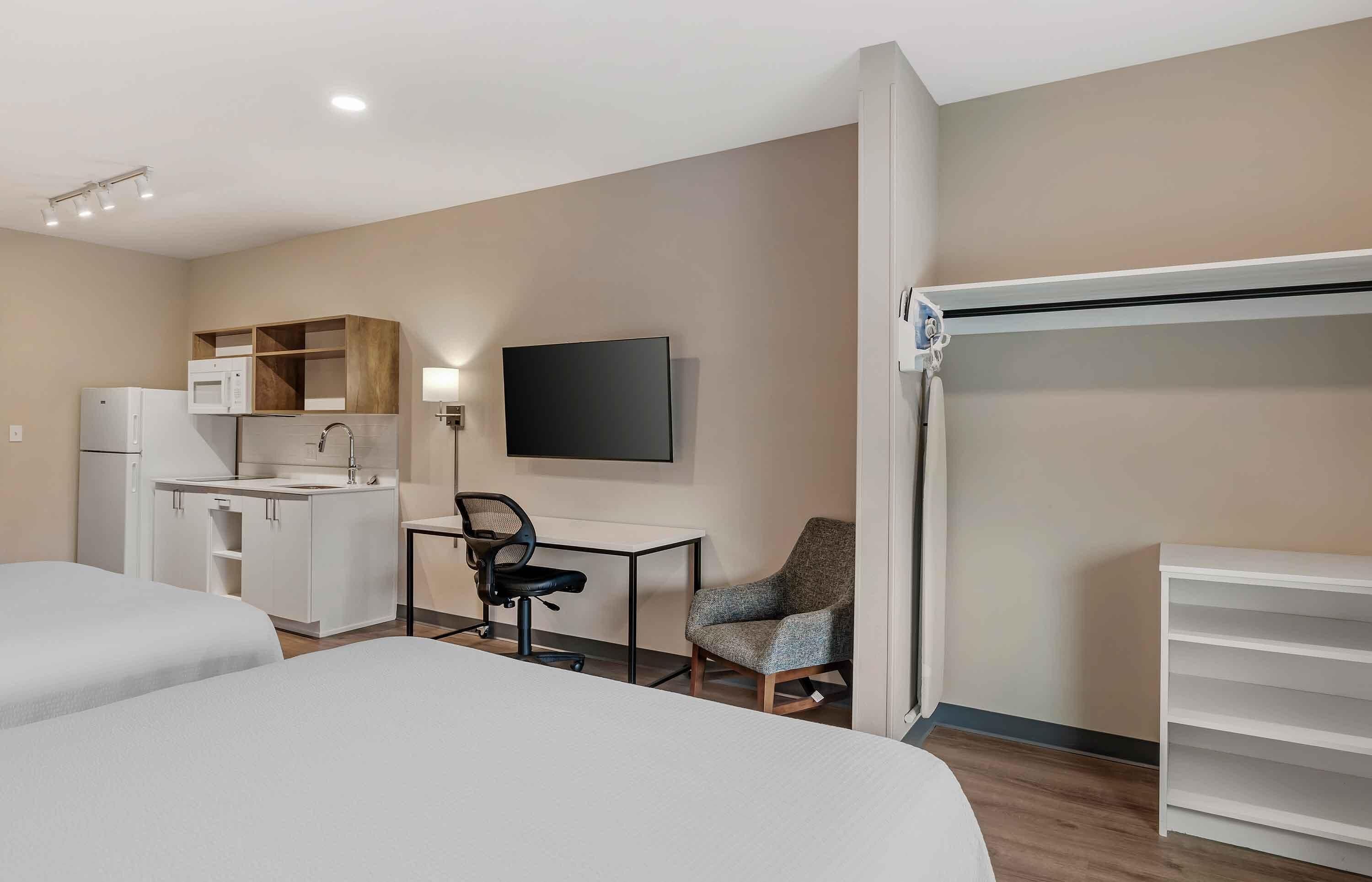 A hotel room with two beds, a kitchenette, a desk with a chair, a TV, and a wardrobe.