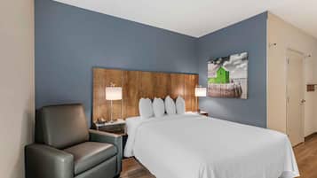A hotel room with a large bed, a wooden headboard, a gray armchair, bedside tables with lamps, and a painting on the wall.